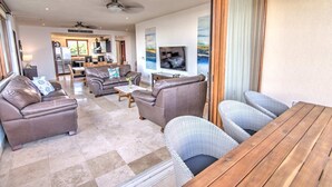 A spacious living area with leather sofas, a wooden table, and wicker chairs. A kitchen is visible in the background.