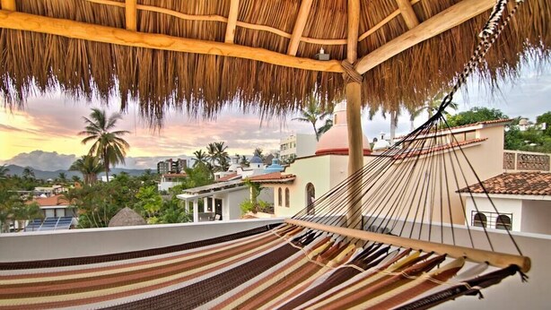 A hammock under a thatched roof, a view of buildings, and palm trees. - Golden Zone Studio • Walk to Beach • Pool • Palapa (Bucerias)