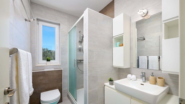 A modern bathroom with a glass shower enclosure, a toilet, a sink with toiletries, and a window with a view. - Villa Galleon with Pool (Murter)