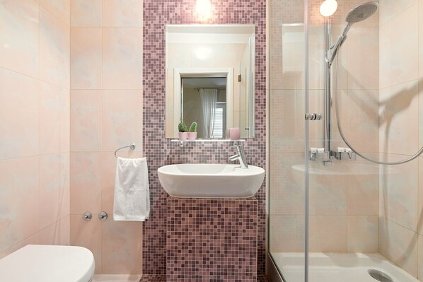 Ein Badezimmer mit einem weißen Waschbecken, einer Duschkabine aus Glas und einer gefliesten Wand mit einem rosa Mosaikmuster.