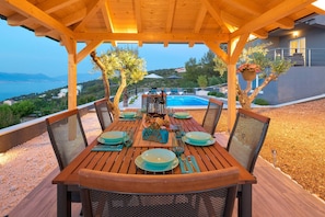 An outdoor dining area with a wooden pergola, a pool in the background, and a view of the sea and hills.