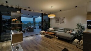 A modern living room with a grey sofa, wooden coffee table, and a dining area with a wooden counter. There is a large window offering a view of the outdoors. - Lovely home in Slivno with WiFi (Slivno)