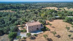 Un bâtiment en pierre de deux étages, surmonté d’un toit en tuiles, entouré d’un paysage aride parsemé d’arbres.