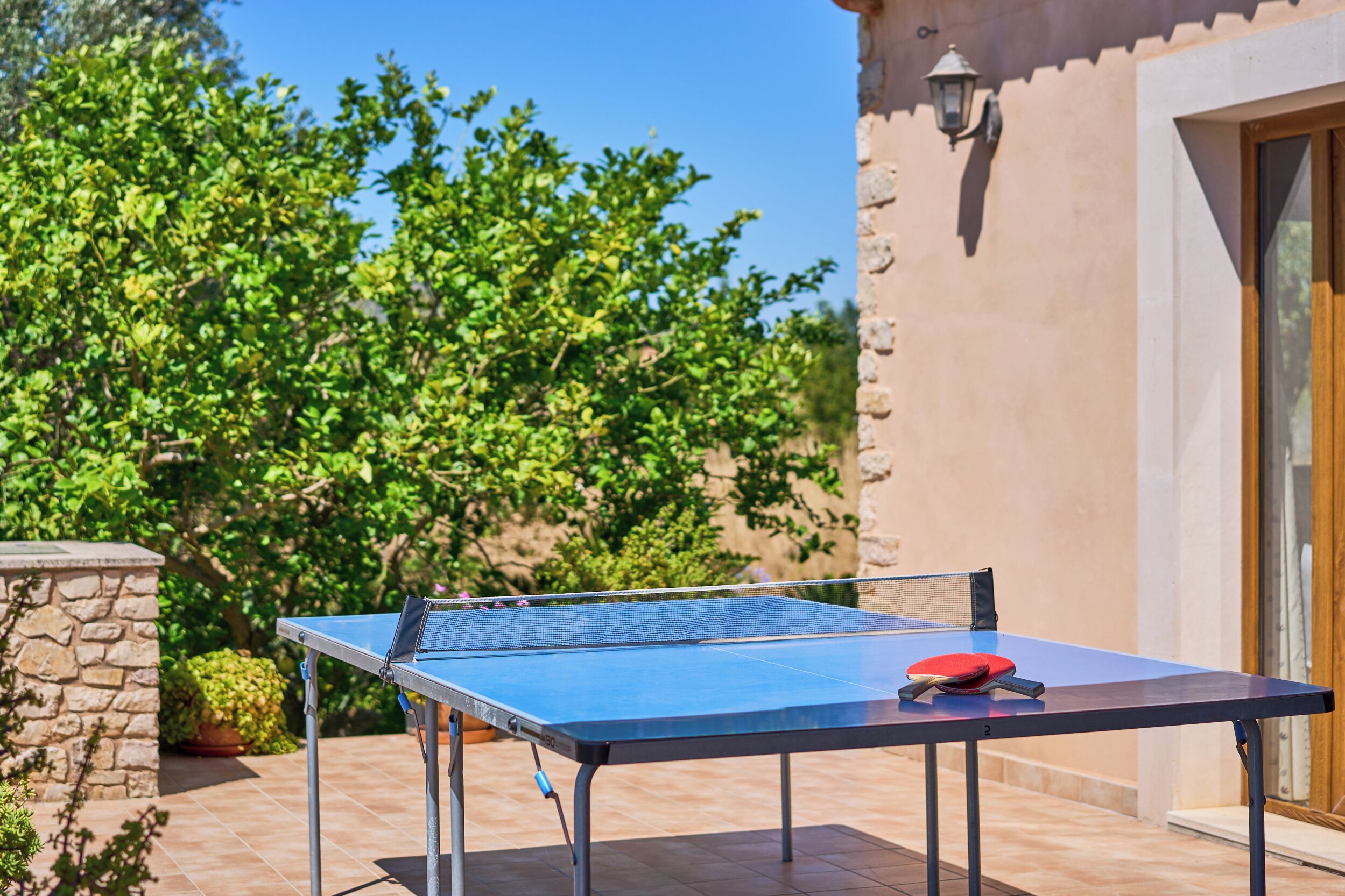 An outdoor ping pong table with a net, a red paddle, and a blue paddle, set against a stone wall and surrounded by greenery.