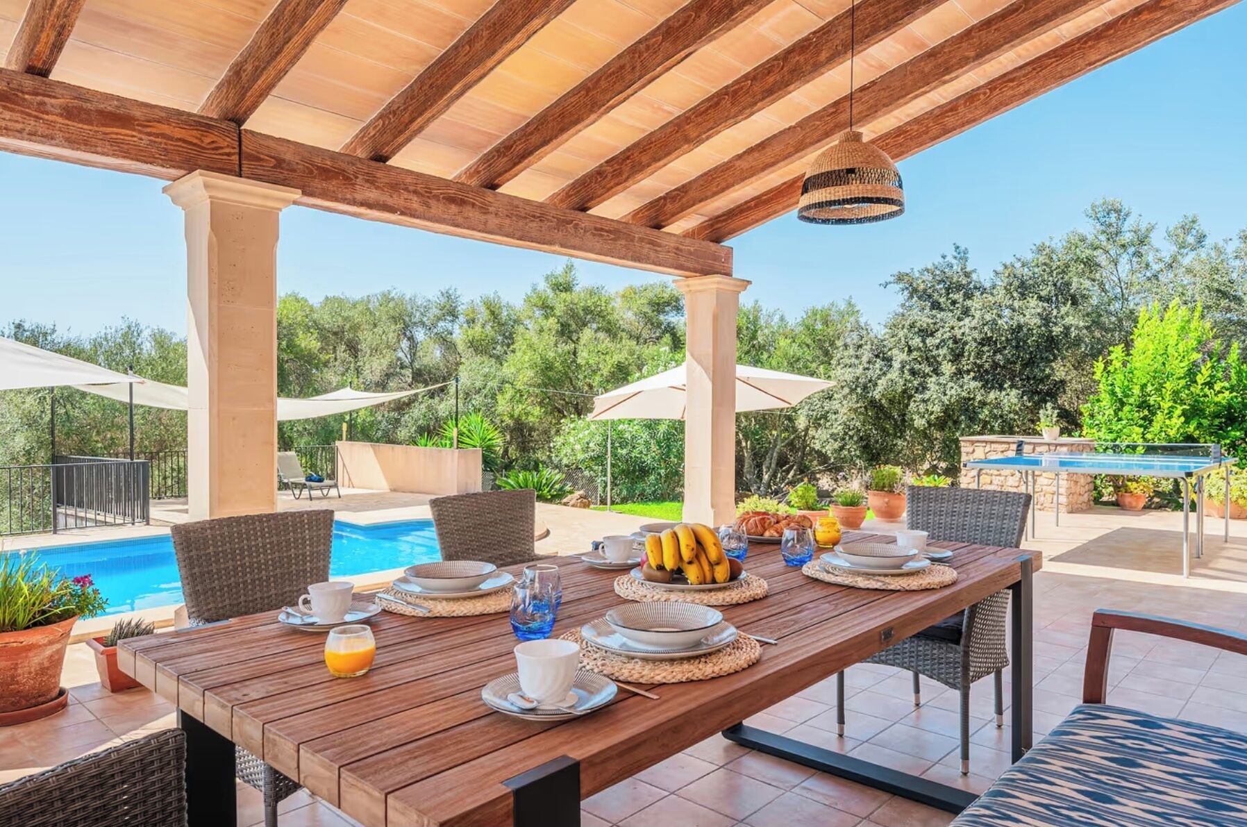 Un espacio exterior con mesas para comer, con una mesa de madera dispuesta para el desayuno, rodeada de sillas de mimbre, cerca de una piscina y cubierto por una pérgola de madera.