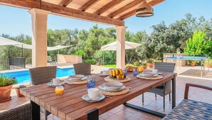Un espace repas extérieur avec une table en bois dressée pour le petit-déjeuner, entourée de chaises en osier, situé près d’une piscine et abrité par une pergola en bois.