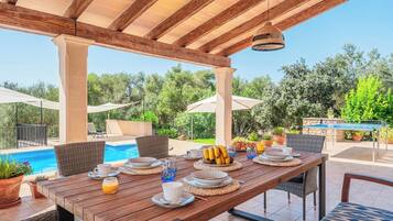 Un espacio exterior con mesas para comer, con una mesa de madera dispuesta para el desayuno, rodeada de sillas de mimbre, cerca de una piscina y cubierto por una pérgola de madera.