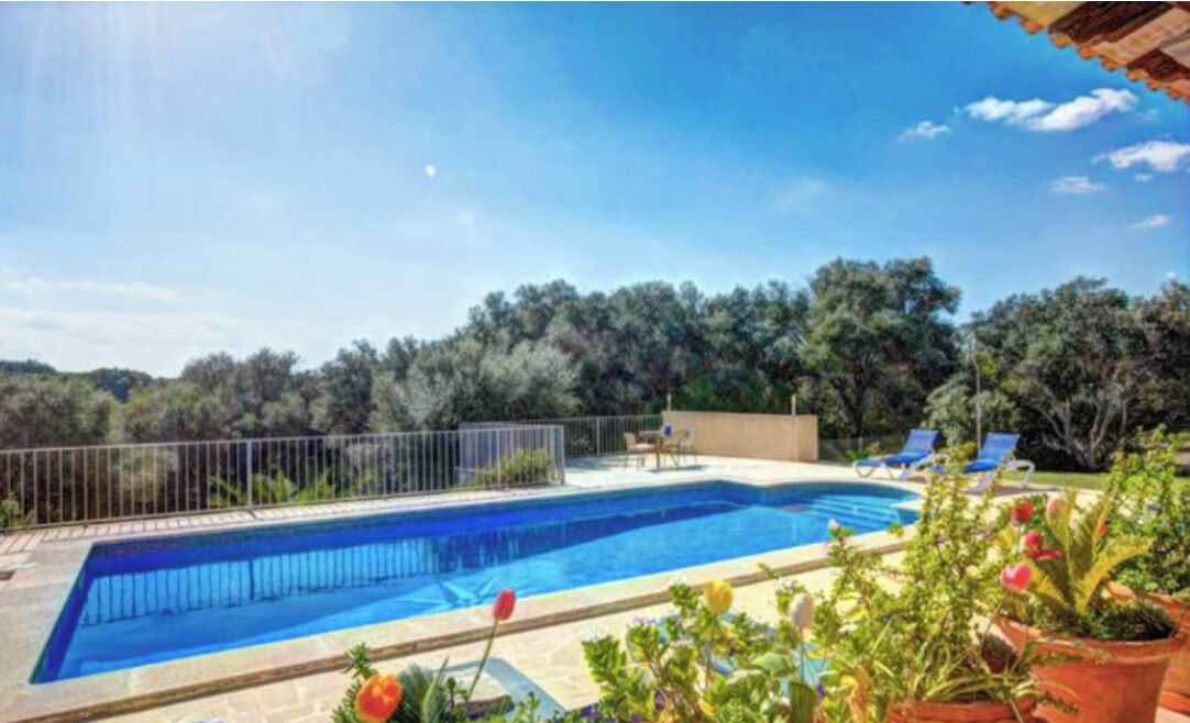 A rectangular swimming pool with a tiled deck, surrounded by a metal fence. There are lounge chairs and a small table on the deck.