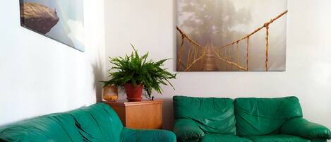 Una sala de estar con dos sofás verdes, un taburete de madera, una planta en maceta y un cuadro de un puente.