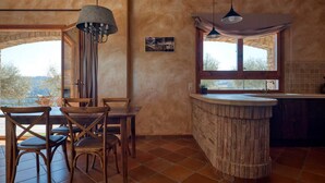 Een rustiek interieur met een houten eettafel en stoelen, een stenen bargedeelte en een groot raam met uitzicht op de omgeving.
