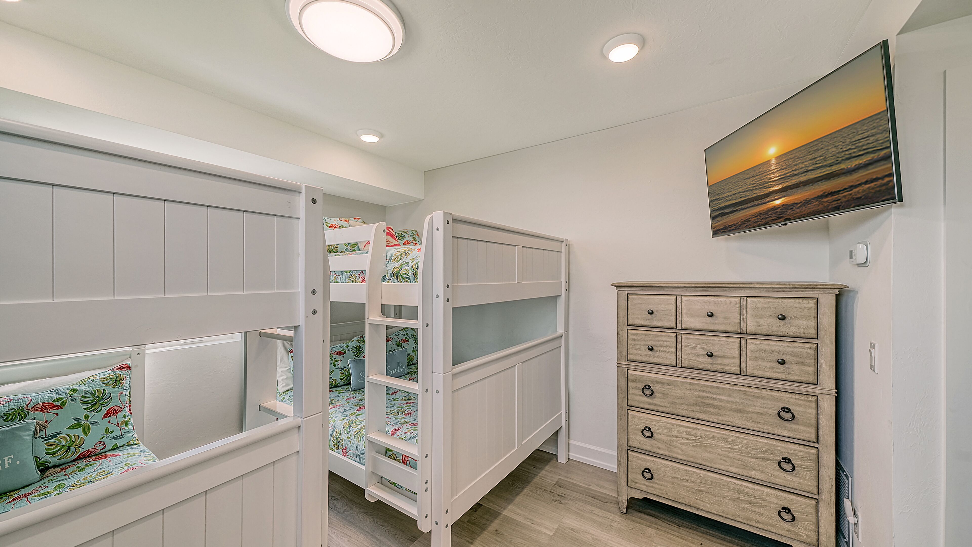A child's bunk bed with floral bedding, a dresser with multiple drawers, and a painting of a sunset on the wall.