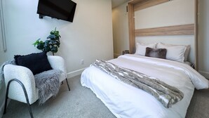 A modern bedroom with a large bed, a mounted TV, a fluffy chair, and a potted plant.