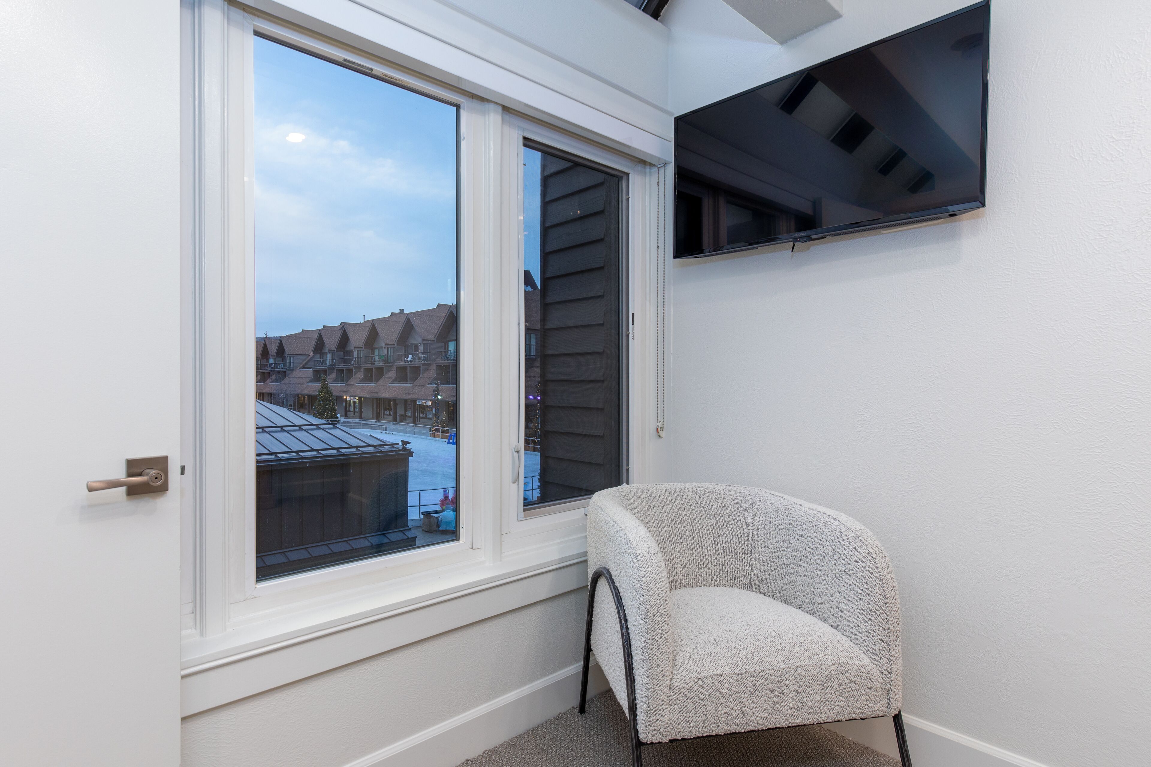 A room with a large window offering a view of neighboring buildings, a white chair, and a mounted flat-screen TV.