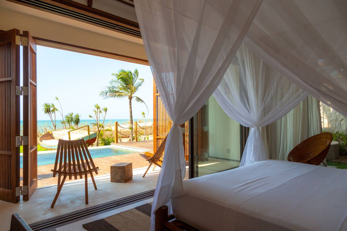 Un dormitorio con vista a la playa, una silla de madera y una cama con ropa blanca.