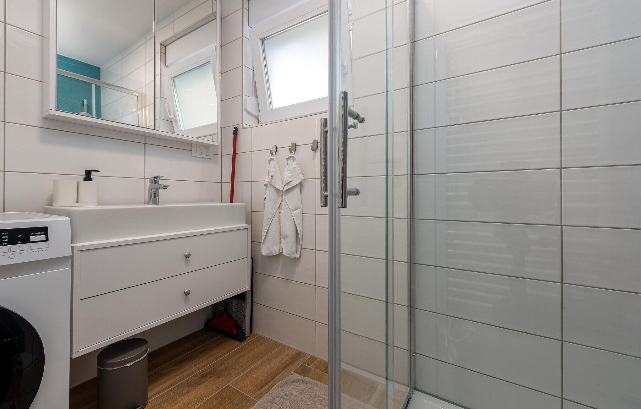 Un bagno moderno con box doccia in vetro, un lavandino bianco con rubinetto, uno specchio sopra il lavandino e un piccolo cestino.