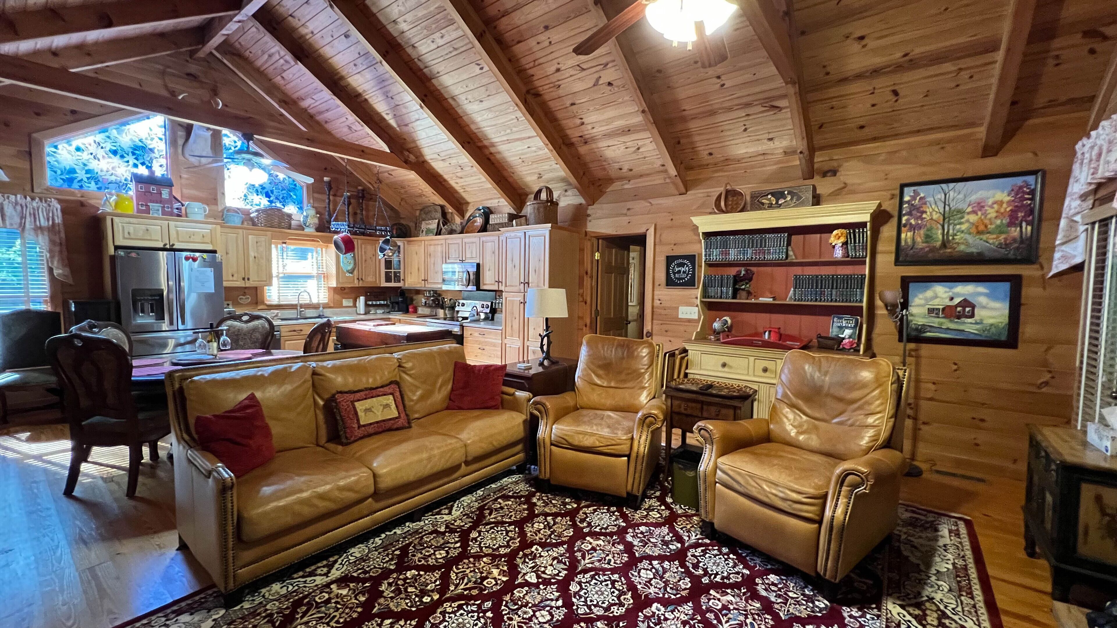 A cozy cabin interior with a wooden ceiling, a dining area, a kitchen, and a living room with leather furniture and a patterned rug.