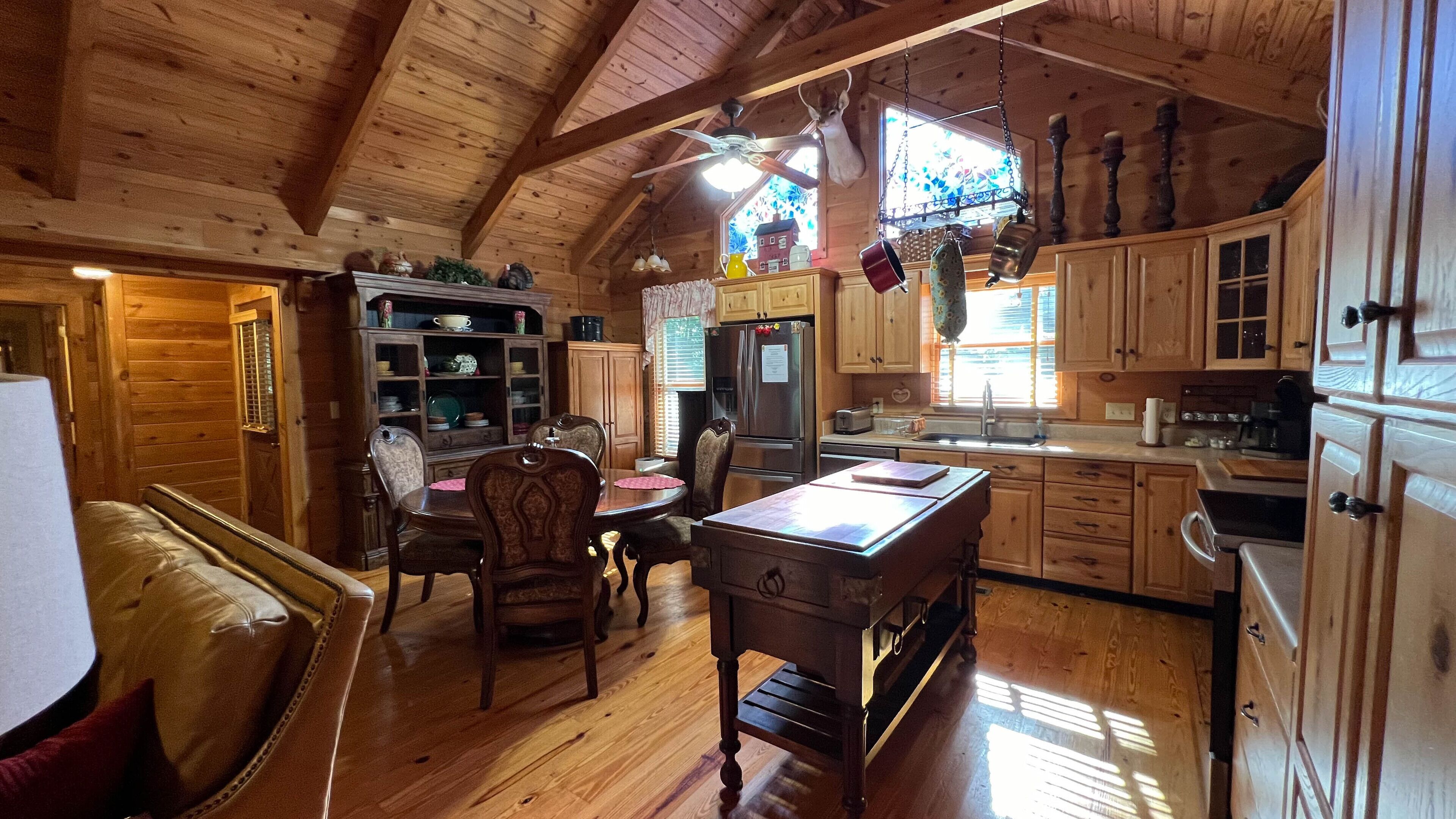 A cozy cabin interior with wooden walls and a high ceiling. It features a dining area with a table and chairs, a kitchen with wooden cabinets and a refrigerator, and a window with a view of a house.