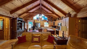 A cozy cabin interior with a high ceiling, wooden beams, a dining area with a leather couch, and a kitchen with wooden cabinets and a large window.