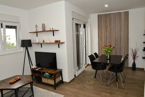 Uma sala de estar moderna com televisão, mesa de centro de madeira e área de jantar com mesa e cadeiras. Há estantes com objetos decorativos e uma porta de vidro deslizante que dá para outro cômodo.