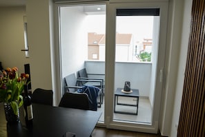 A modern living room with a dining table, chairs, a sofa, and a small table with a decorative item. A sliding glass door leads to an outdoor balcony with a view of buildings. - Apartment Nicole - One Bedroom Apartment with Balcony (Zadar)