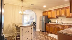 A kitchen with wooden cabinets, a stainless steel refrigerator, and a dining area with a white table and chairs. - Full Kitchen // Spacious Living Room // Walk to Mineral Springs and Restaurants (Hot Springs)