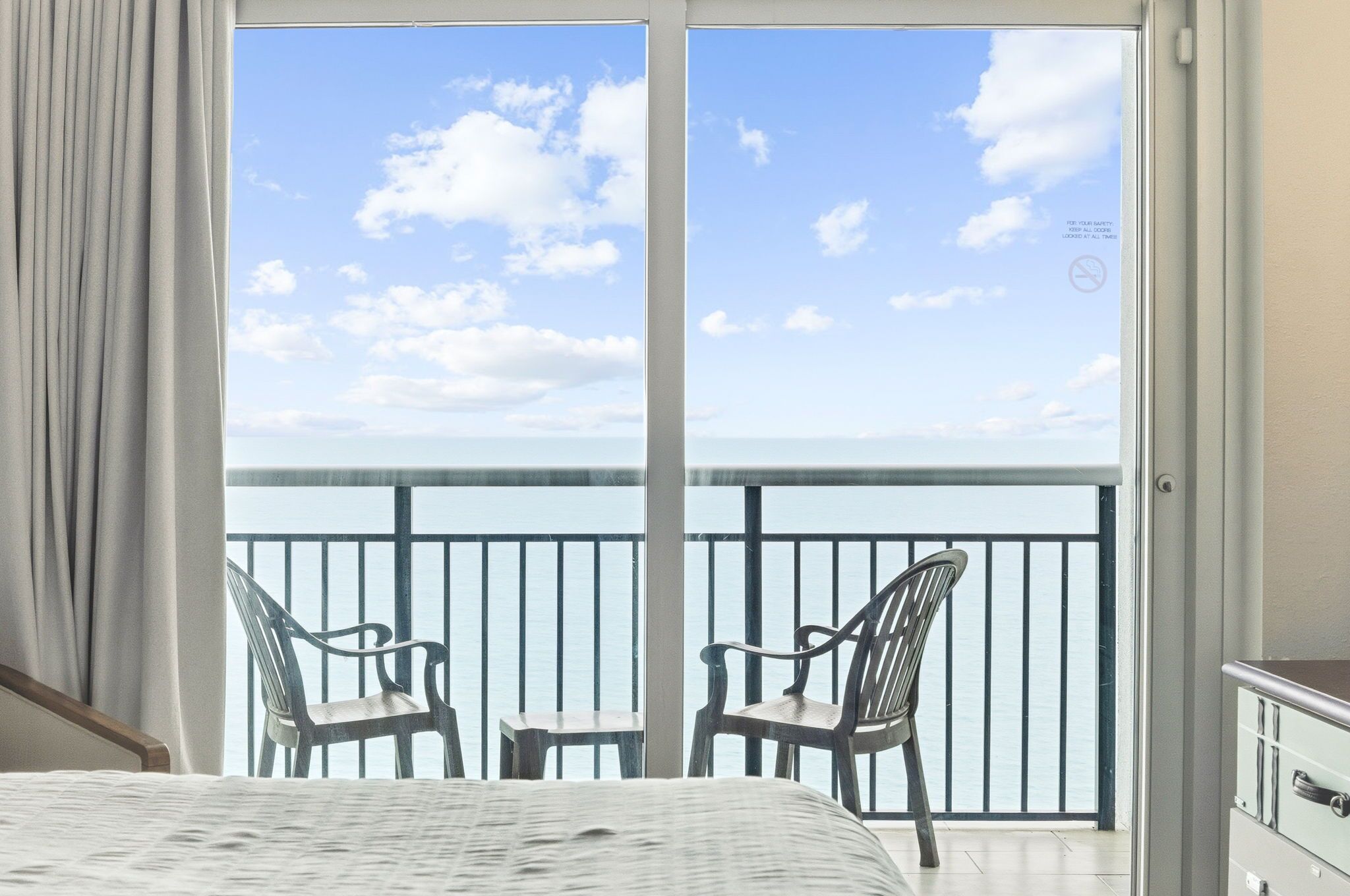 A room with a bed, two chairs, and a view of the sea through a balcony door.