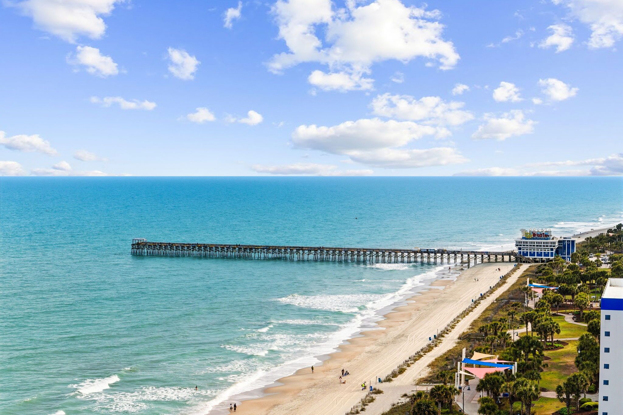Ein Strand mit einem Steg, der ins Meer führt, ein Gebäude mit blauem Dach und ein klarer Himmel.