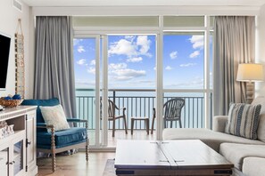 A room with a sofa, a chair, a coffee table, a TV, and a balcony with a view of the sky and clouds.