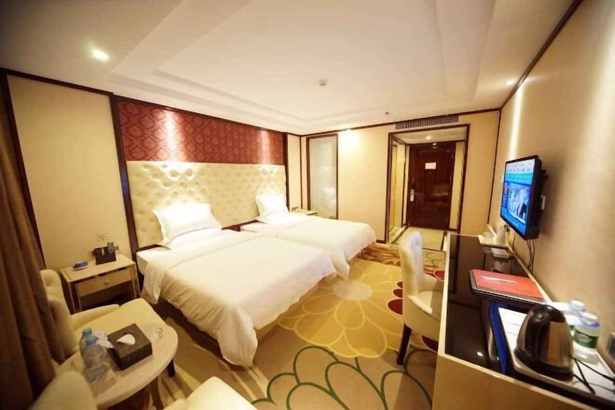 Quarto de hotel com duas camas, uma escrivaninha com computador, uma cadeira, uma mesinha com uma garrafa e uma televisão na parede.