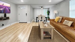 A modern living room with a flat-screen TV, a brown leather sofa, a wooden coffee table, and a dining area with a white table and chairs. - La Casita of St. Petersburg near Beaches and Bars! (St. Petersburg)