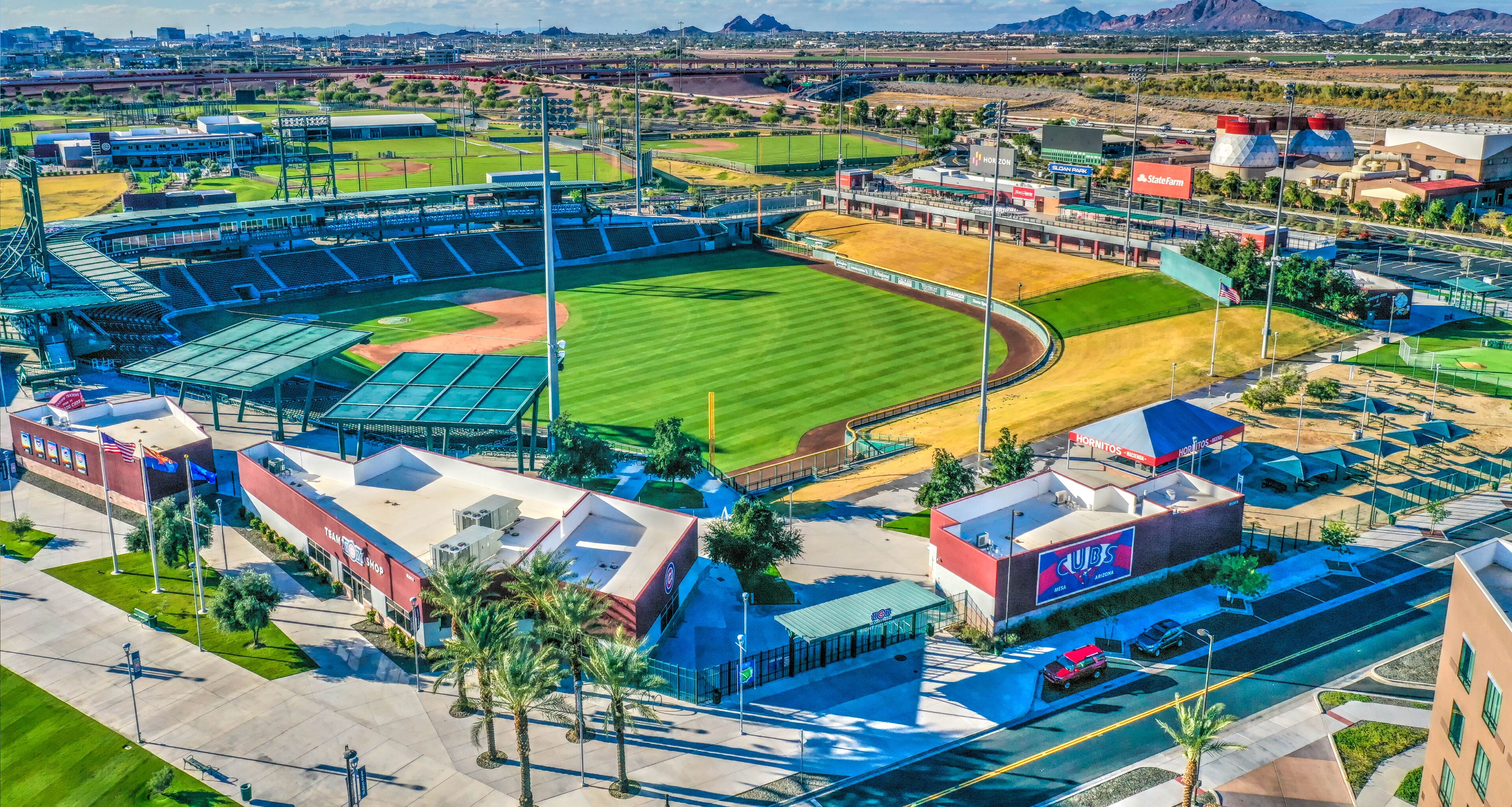 An aerial view of a baseball stadium with a large seating area, a field, and surrounding buildings including a hotel and shops.
