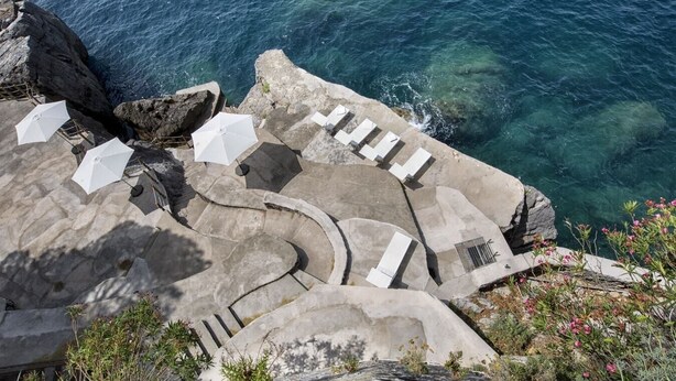 A stone terrace with white umbrellas and lounge chairs overlooking a clear blue sea.