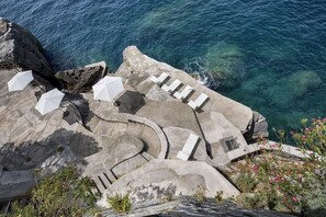 Una terrazza in pietra con ombrelloni bianchi e poltrone prendisole affacciata su un mare cristallino e blu.