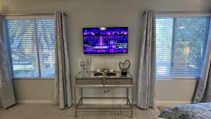 Een slaapkamer met een aan de muur bevestigde televisie, een nachtkastje met decoratieve objecten, en een bed met een bloemenprint beddekleed.