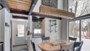 A dining area with a wooden table and chairs, a kitchen in the background, and a large window with a view of trees. - Forest Chalet Just 5 min to Mt Orford Avail August - December 2026 (Orford)