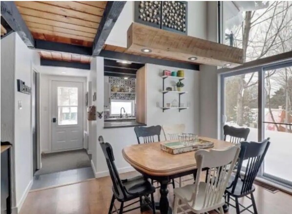 Enjoy meals in this spacious dining area with a wooden table and four chairs, surrounded by a high ceiling and large window offering a view of the outdoors.