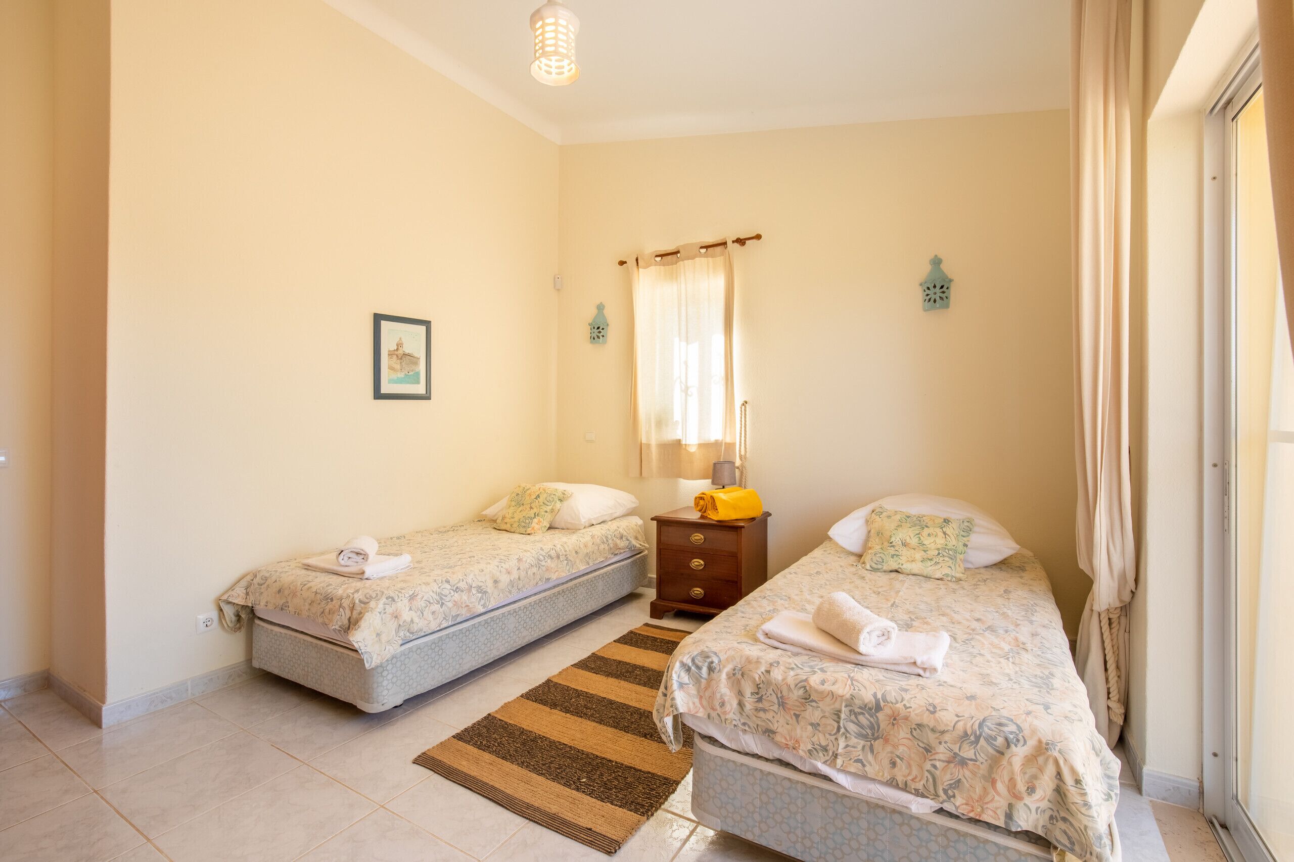 A bedroom with two beds, a bedside table with a lamp, a window with curtains, and a small wall decoration.