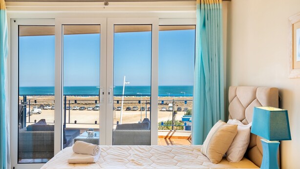 A hotel room with a bed, a nightstand, a lamp, and a view of the beach and ocean through large sliding doors. - Casa Areia Ornata, sea view terrace, beach (Lagos)