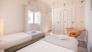 Quarto de hotel com duas camas, uma janela com cortinas, uma luminária na parede e uma cadeira de madeira com toalhas.