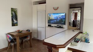 Ett kompakt kök med ett kylskåp, ett matbord med stolar, en TV som är monterad på väggen och en liten bardisk med flaskor.