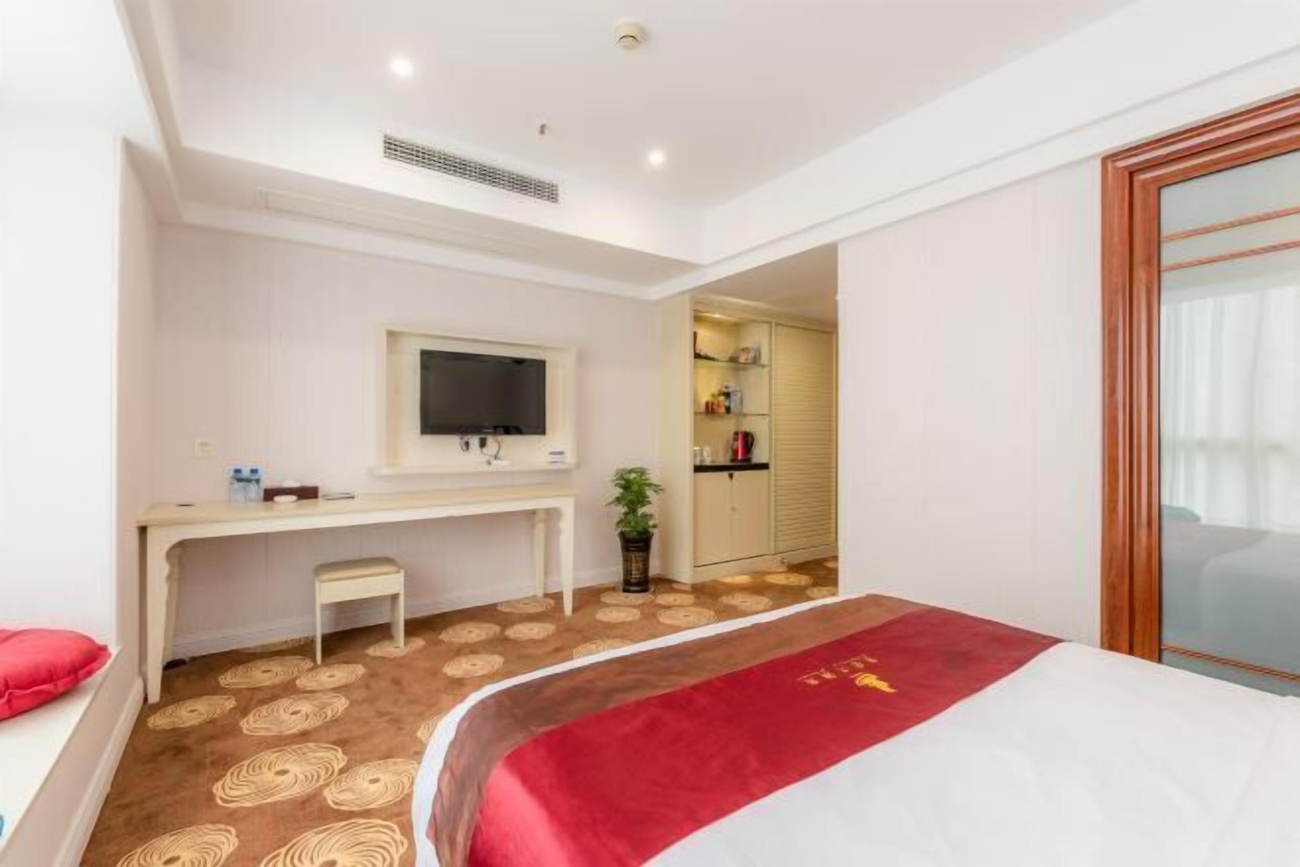 A hotel room with a bed, a desk, a television, a small table, a plant, and a kitchenette.