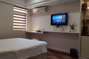 Een hotelkamer met een bed, een tv met ingebouwde bevestiging waarop een videogesprek wordt weergegeven, een klein plankje met spullen, en een raam met jaloezieën.