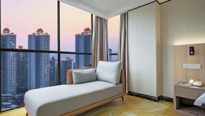 Una habitación de hotel moderna con un ventanal grande, un sofá cama, una mesita de noche con teléfono y una lámpara de pared.