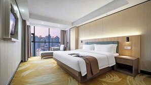 Habitación de hotel con una cama grande, una mesita de noche, un televisor montado en la pared y vistas a la ciudad a través de amplios ventanales.