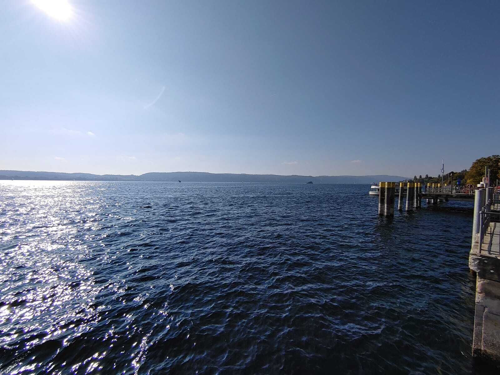 Vista al agua con un muelle que se adentra en el agua, cielo despejado y formas de tierra a lo lejos.