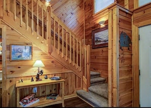 A wooden interior with a staircase, a framed picture, a table with a lamp, and a small shelf with figurines. - Hottub* Great Location 3.5 Miles Gatlinburg (Gatlinburg)