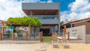 A modern building with a flat roof, a covered outdoor seating area with chairs, and a sandy ground.