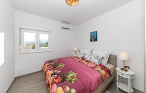 Un dormitorio con cama, ventana, aire acondicionado, mesita de noche y lámpara.