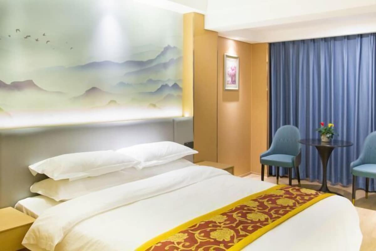 A hotel room with a large bed, two chairs, a small table with a vase, and a painting of mountains on the wall.