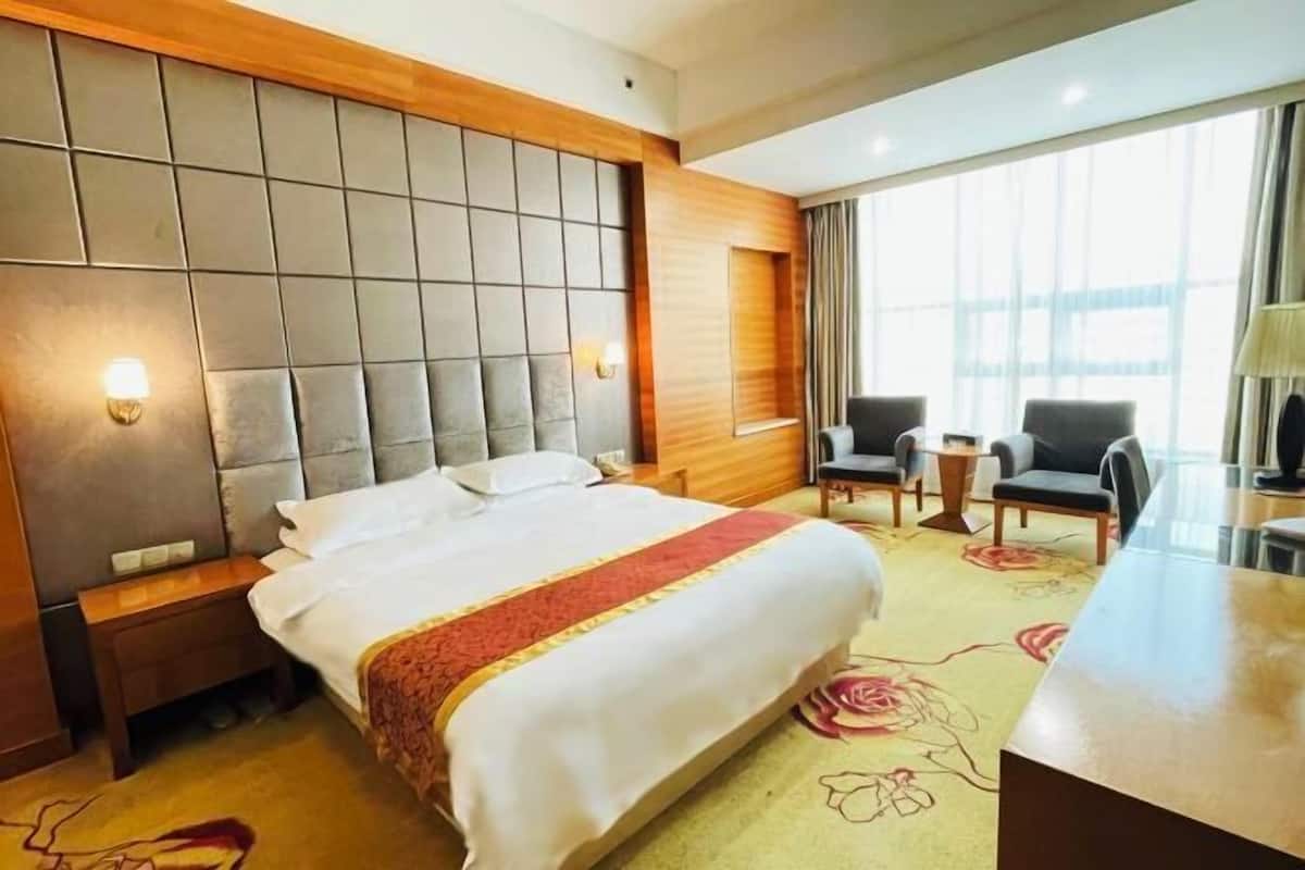 Habitación de hotel con una cama grande, cabecera de madera, una mesita, dos sillas y una ventana con cortinas.